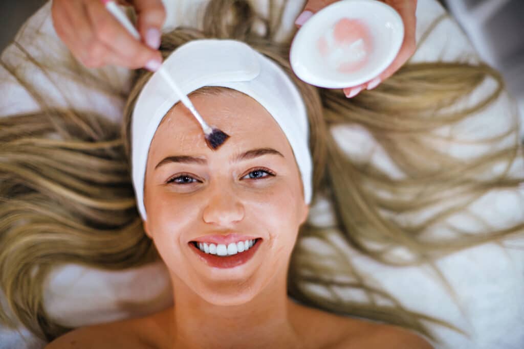 Woman receiving a facial chemical peel treatment as a provider applies the solution to her forehead. Woman receiving a facial chemical peel treatment as a provider applies the solution to her forehead.