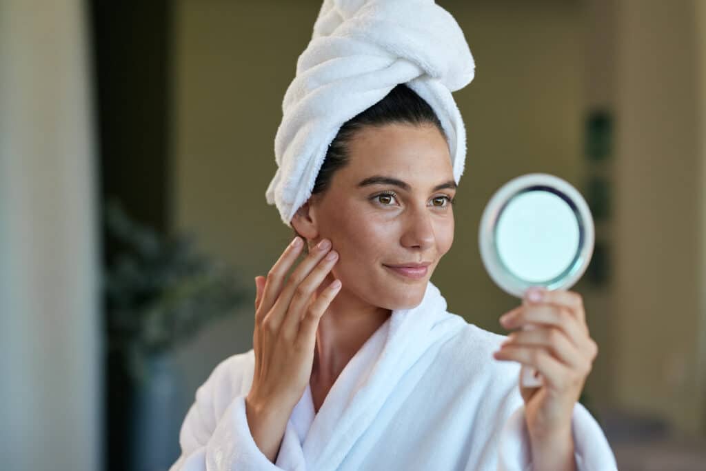 Woman in a bathrobe examines her facial skin in a handheld mirror after a skin care routine. Woman in a bathrobe examines her facial skin in a handheld mirror after a skin care routine.